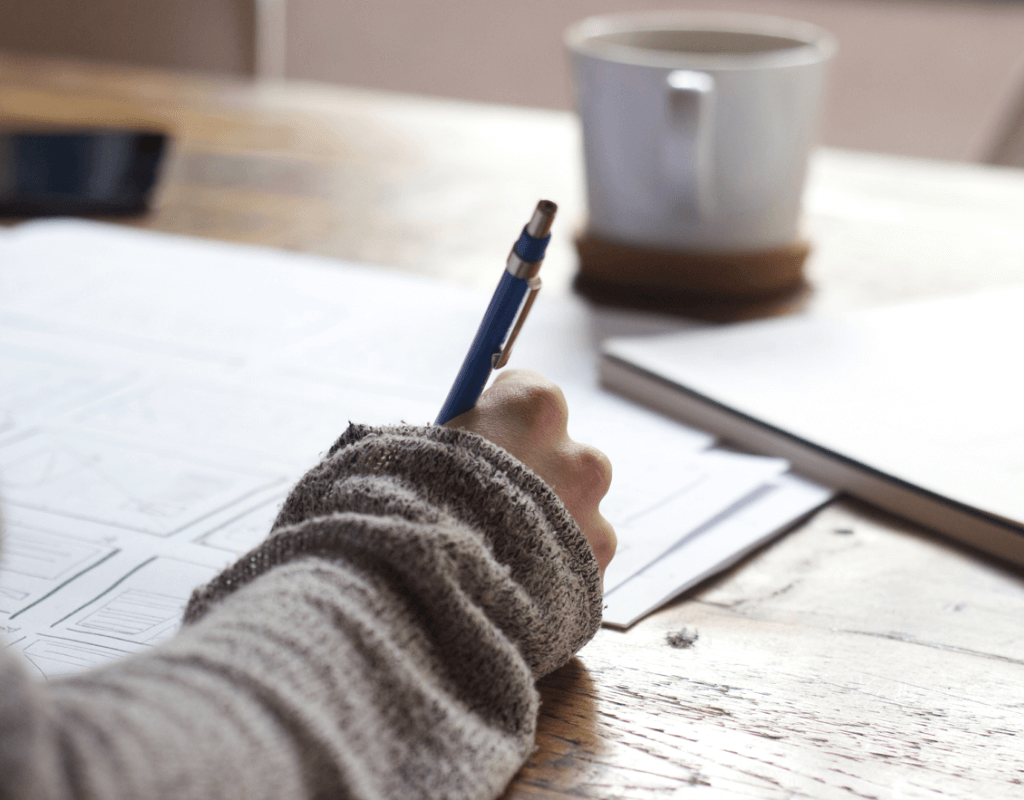someone journaling on paper. Shown is the arm, paper journal and a cup sitting on a table